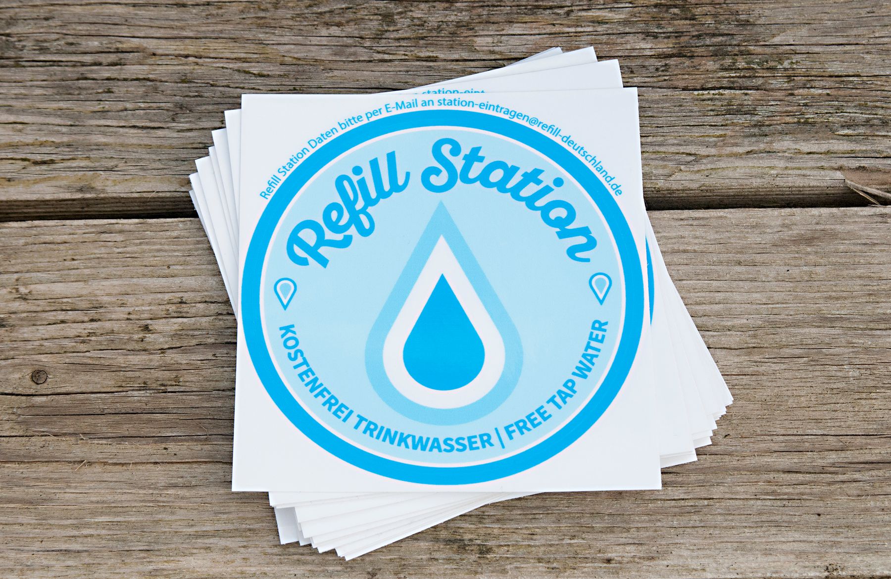 A stack of blue and white stickers reads Refill Station with a water drop icon and the text Kostenfrei Trinkwasser / Free Tap Water on a wooden surface.
